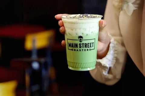 Main Street Roasters honeycomb matcha latte held by hand in a clear cup, featuring layered green matcha, creamy foam topping, and the café logo on the front