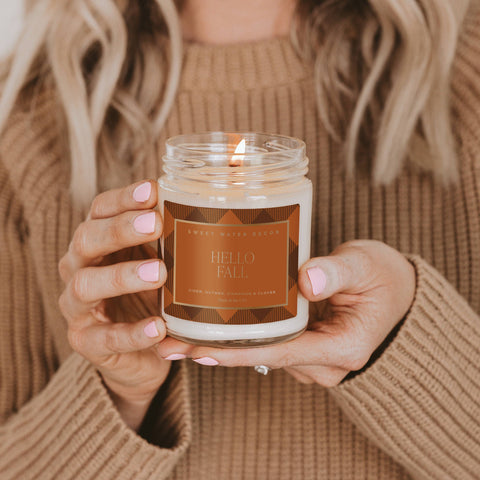 Person holding a 'Hello Fall' candle with a warm, autumnal design.