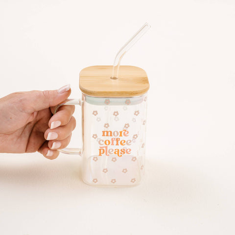 A hand with manicured nails holds a clear glass mug with a bamboo lid and a clear glass straw. The mug is decorated with small pink floral patterns and the text 'more coffee please' in orange.