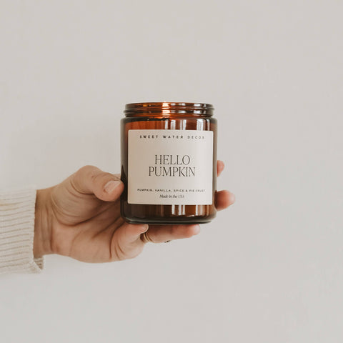 Hand holding a candle labeled 'Hello Pumpkin' by Sweet Water Decor against a plain background