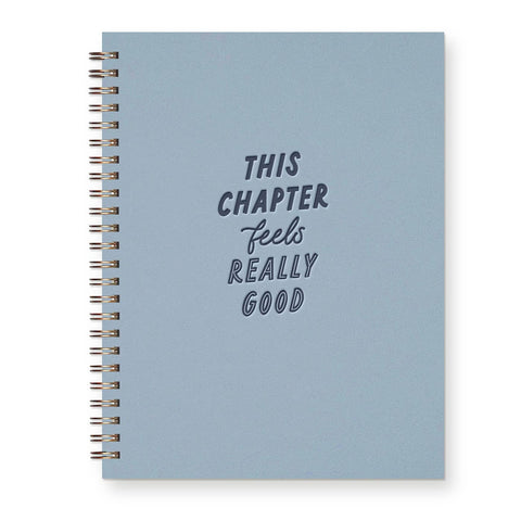 A light blue spiral notebook with a silver wire binding and the text 'THIS CHAPTER feels REALLY GOOD' printed on the front cover, shown against a plain white background.