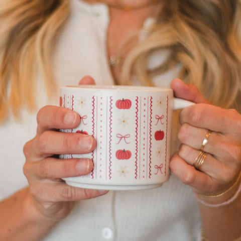 A person holding a white ceramic mug with a red and pink autumn pattern of pumpkins, bows, and zigzags, wearing a white ribbed top and gold rings.