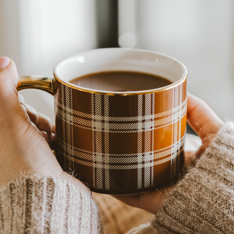 Person holding a plaid mug with coffee in a cozy setting