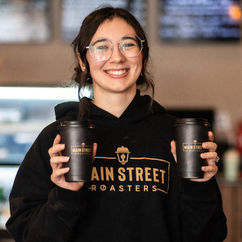Person wearing a 'Main Street Roasters' hoodie holding two coffee cups in a coffee shop.