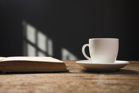 White coffee cup on a saucer next to an open book on a wooden table with a dark background