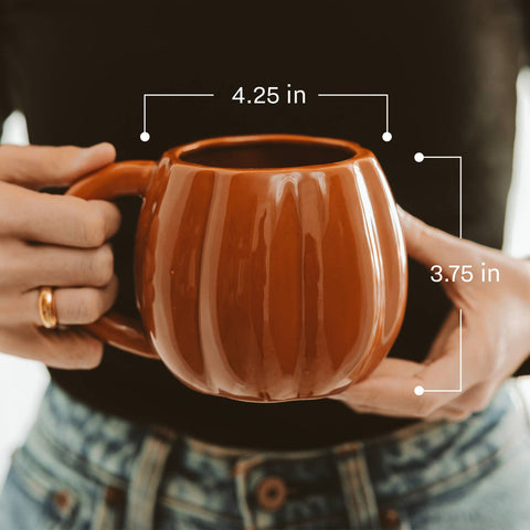 Brown ceramic mug held by hands with measurements indicated, against a blurred background.