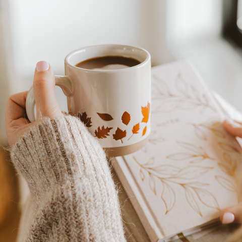 Person holding a mug with a leaf pattern next to a book on a light surface.