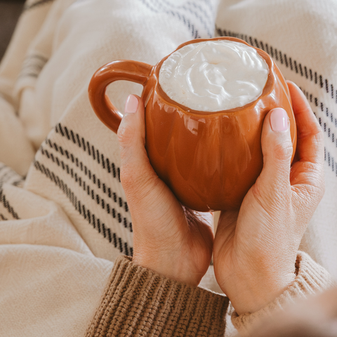 Person holding a pumpkin mug with a hot beverage and whipped cream, wearing a beige sweater.