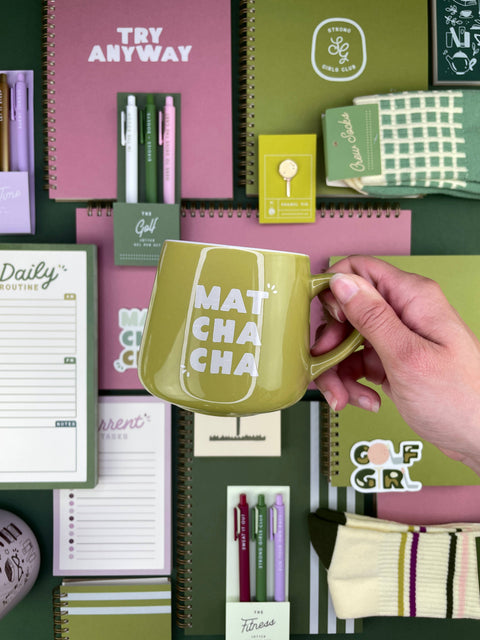 A hand holds a green ceramic mug with the words 'MATH CHA CHA' printed in white. The mug is surrounded by coordinated stationery items including notebooks, pens, sticky notes, and a sock on a flat-lay background.