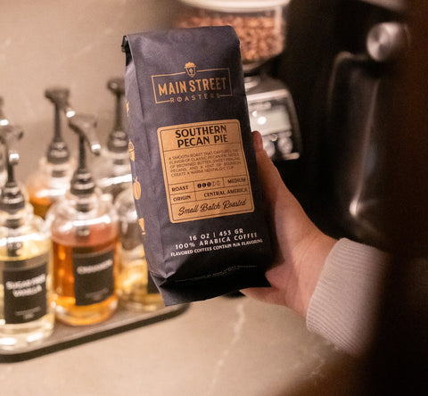 Hand holding a package of Main Street Coffee in a coffee shop setting with coffee beans and bottles in the background.