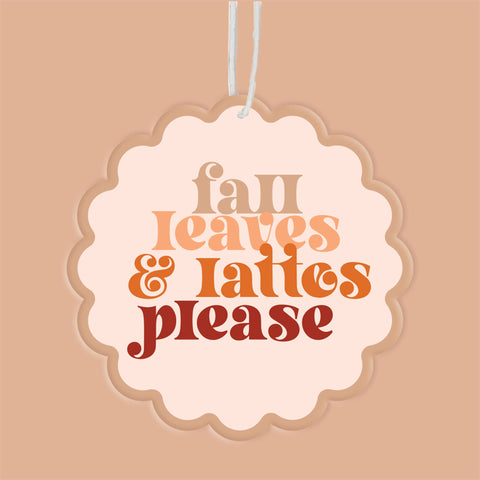 A scalloped oval hanging air freshener with the phrase 'fall leaves & lattes please' in decorative autumn-colored lettering, suspended by a thin white string against a warm beige background.