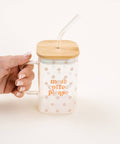 A hand with manicured nails holds a clear glass mug with a bamboo lid and a clear glass straw. The mug is decorated with small pink floral patterns and the text 'more coffee please' in orange.
