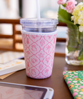 Pink patterned cup sleeve on a clear  with a straw on a table with flowers and stationery in the background