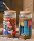 Two clear glass tumblers with bamboo lids and metal straws filled with iced coffee, decorated with colorful illustrations. One tumbler features book-themed graphics like a ballet shoe and library card, and the other features a city skyline with a taxi.