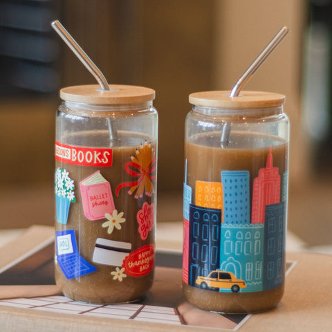 Two clear glass tumblers with bamboo lids and metal straws filled with iced coffee, decorated with colorful illustrations. One tumbler features book-themed graphics like a ballet shoe and library card, and the other features a city skyline with a taxi.
