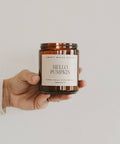Hand holding a candle labeled 'Hello Pumpkin' by Sweet Water Decor against a plain background