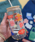A hand holds a clear glass jar tumbler with a bamboo lid and metal straw. The glass features colorful printed stickers and text including 'BOOKS', 'BILLET shop', 'shop girl', and 'HAPPY thanksgiving BACK'.