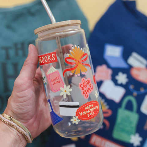 A hand holds a clear glass jar tumbler with a bamboo lid and metal straw. The glass features colorful printed stickers and text including 'BOOKS', 'BILLET shop', 'shop girl', and 'HAPPY thanksgiving BACK'.