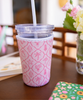 Pink patterned cup sleeve on a clear  with a straw on a table with flowers and stationery in the background