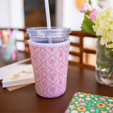 Pink patterned cup sleeve on a clear  with a straw on a table with flowers and stationery in the background