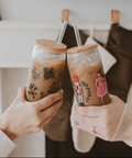 Two hands holding iced coffee drinks with festive designs against a blurred indoor background.