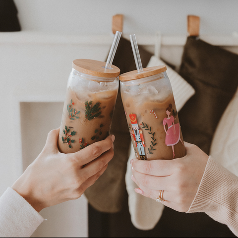 Two hands holding iced coffee drinks with festive designs against a blurred indoor background.