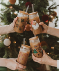 Four hands holding festive-themed iced drinks in front of a decorated Christmas tree.