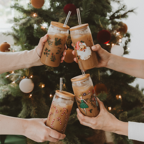 Four hands holding festive-themed iced drinks in front of a decorated Christmas tree.