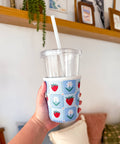 Hand holding a clear cup with a colorful strawberry pattern sleeve, against a home interior background.
