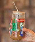 A hand holding a clear glass jar tumbler with a bamboo lid and metal straw, filled with a brown beverage. The jar features colorful holiday-themed illustrations and a red 'FOX & SONS' label.