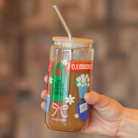 A hand holding a clear glass jar tumbler with a bamboo lid and metal straw, filled with a brown beverage. The jar features colorful holiday-themed illustrations and a red 'FOX & SONS' label.