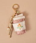 A coffee cup-shaped keychain bag in dusty pink and white with white polka dots, featuring a printed orange flower illustration on the front. It has a gold metal key ring, a beige braided rope charm with a small pearl bead, and is photographed on a solid beige background.