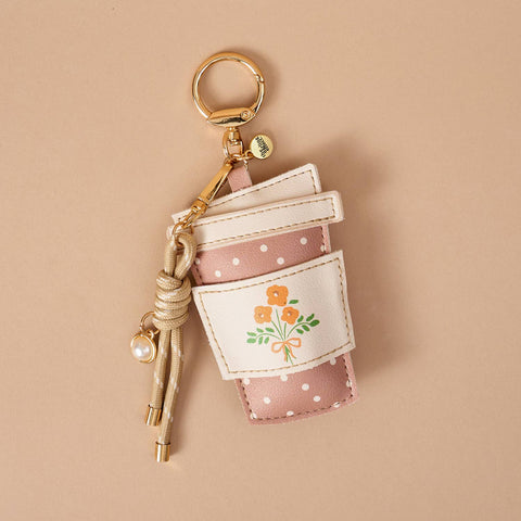 A coffee cup-shaped keychain bag in dusty pink and white with white polka dots, featuring a printed orange flower illustration on the front. It has a gold metal key ring, a beige braided rope charm with a small pearl bead, and is photographed on a solid beige background.