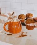 Person holding a muffin next to an pumpkin mug with whipped cream on a kitchen counter.