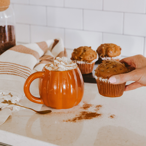 Person holding a muffin next to an pumpkin mug with whipped cream on a kitchen counter.