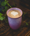 Pink ceramic cup with a heart-shaped foam design on a wooden surface with greenery.