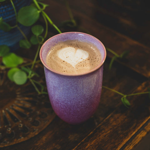 Pink ceramic cup with a heart-shaped foam design on a wooden surface with greenery.