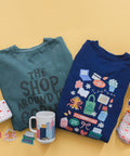 Two folded graphic T-shirts are displayed on a warm yellow background, surrounded by small lifestyle props including a mug, stickers, a cup, and a patterned pouch. The left shirt is teal with dark gray text reading 'THE SHOP AROUND YOU', and the right shirt is navy with colorful illustrated labels and phrases.