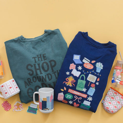 Two folded graphic T-shirts are displayed on a warm yellow background, surrounded by small lifestyle props including a mug, stickers, a cup, and a patterned pouch. The left shirt is teal with dark gray text reading 'THE SHOP AROUND YOU', and the right shirt is navy with colorful illustrated labels and phrases.