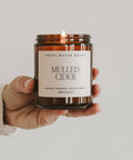 Hand holding a 'Mulled Cider' candle with a label on a neutral background