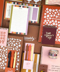 A styled flat-lay collection of stationery items including notebooks, a weekly checklist pad, pens, washi tape, and notepads in coordinated pink, orange, cream, and brown tones on a brown background.