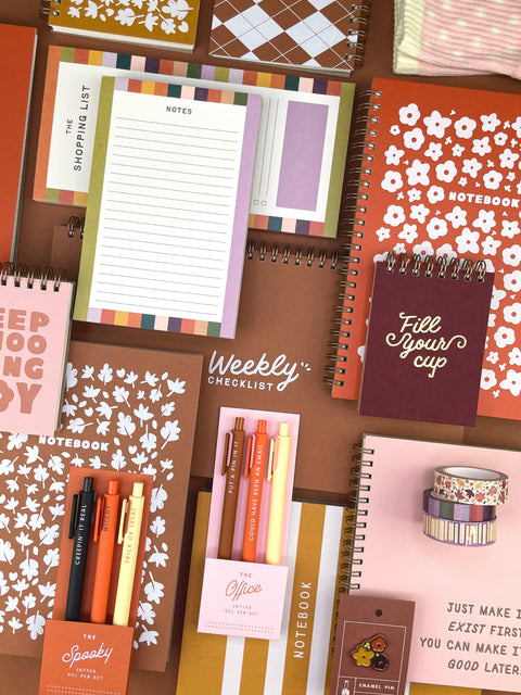 A styled flat-lay collection of stationery items including notebooks, a weekly checklist pad, pens, washi tape, and notepads in coordinated pink, orange, cream, and brown tones on a brown background.