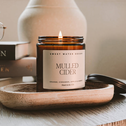 Candle labeled 'Mulled Cider' by Sweet Water Decor on a wooden tray with books and glasses in the background.