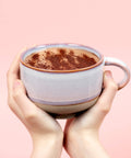 Two hands holding a mug of hot chocolate with cocoa powder on a pink background