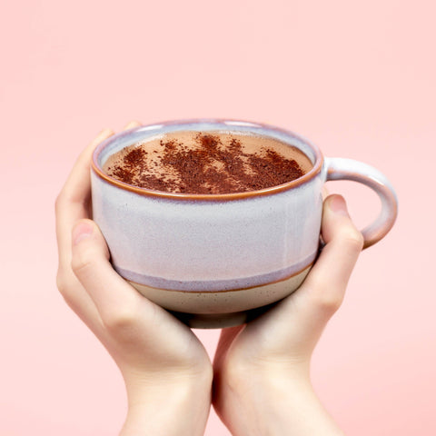 Two hands holding a mug of hot chocolate with cocoa powder on a pink background