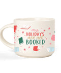 An off-white ceramic mug with a holiday-themed print that reads 'my HOLIDAYS are all BOOKED' in pink and green lettering, surrounded by illustrations of open books and stars.
