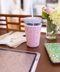 Pink patterned cup sleeve on a clear tumbler with straw on a table with a tablet and flowers in the background