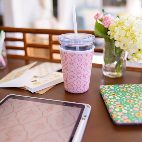 Pink patterned cup sleeve on a clear tumbler with straw on a table with a tablet and flowers in the background