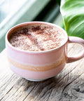 Pink mug with a hot beverage on a wooden surface with green leaves in the background