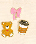Set of three pins including a teddy bear, coffee cup, and pink bow on a beige background